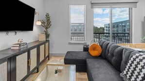 Ein modernes Wohnzimmer mit einer Eckcouch, einem Flachbildfernseher, einem Glastisch und Blick auf die Stadt durch große Fenster.