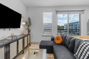 Ein modernes Wohnzimmer mit einer Eckcouch, einem Flachbildfernseher, einem Glastisch und Blick auf die Stadt durch große Fenster.