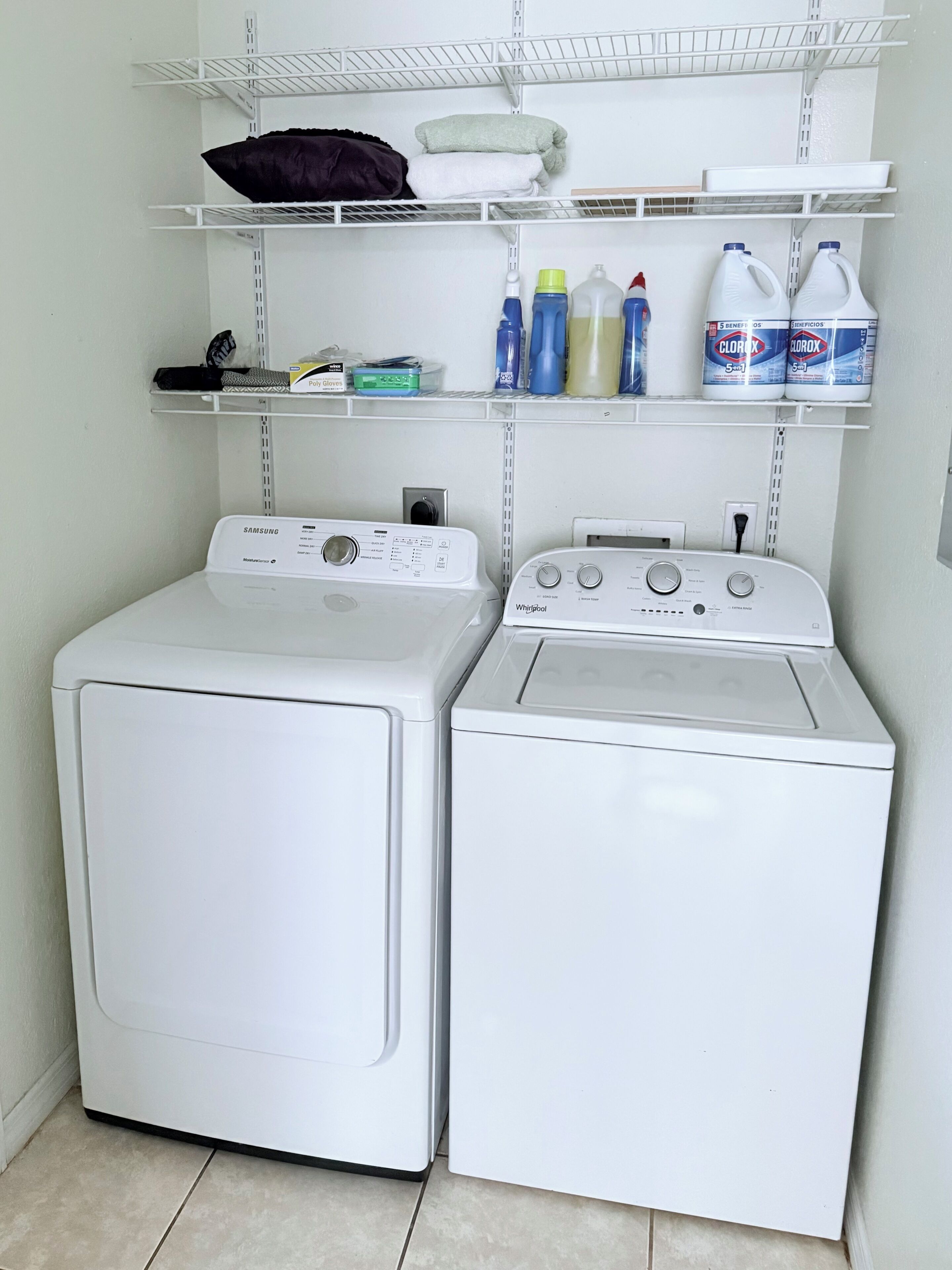 A laundry area with two front-loading washing machines and shelves above containing folded towels and cleaning supplies.