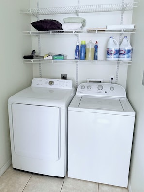 A laundry area with two front-loading washing machines and shelves above containing folded towels and cleaning supplies.