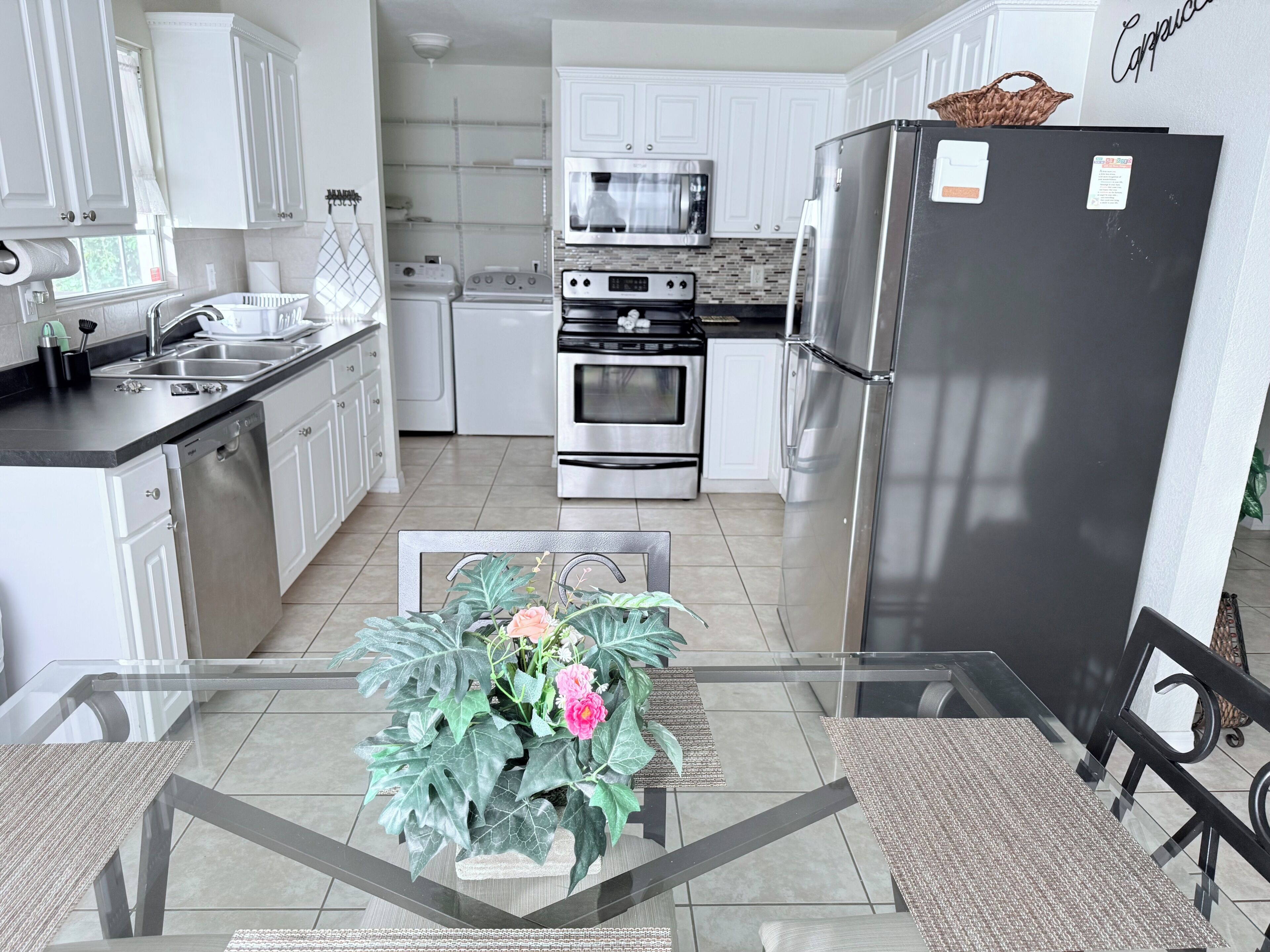 A modern kitchen with white cabinetry, stainless steel appliances, and a glass dining table with a centerpiece.