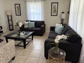 A modern living room with a black sofa, a glass coffee table, and a patterned armchair. There are two framed pictures on the wall, a floor lamp, and a small side table with a plant. - 3-bedroom bungalow in peaceful George Town with AC (George Town)