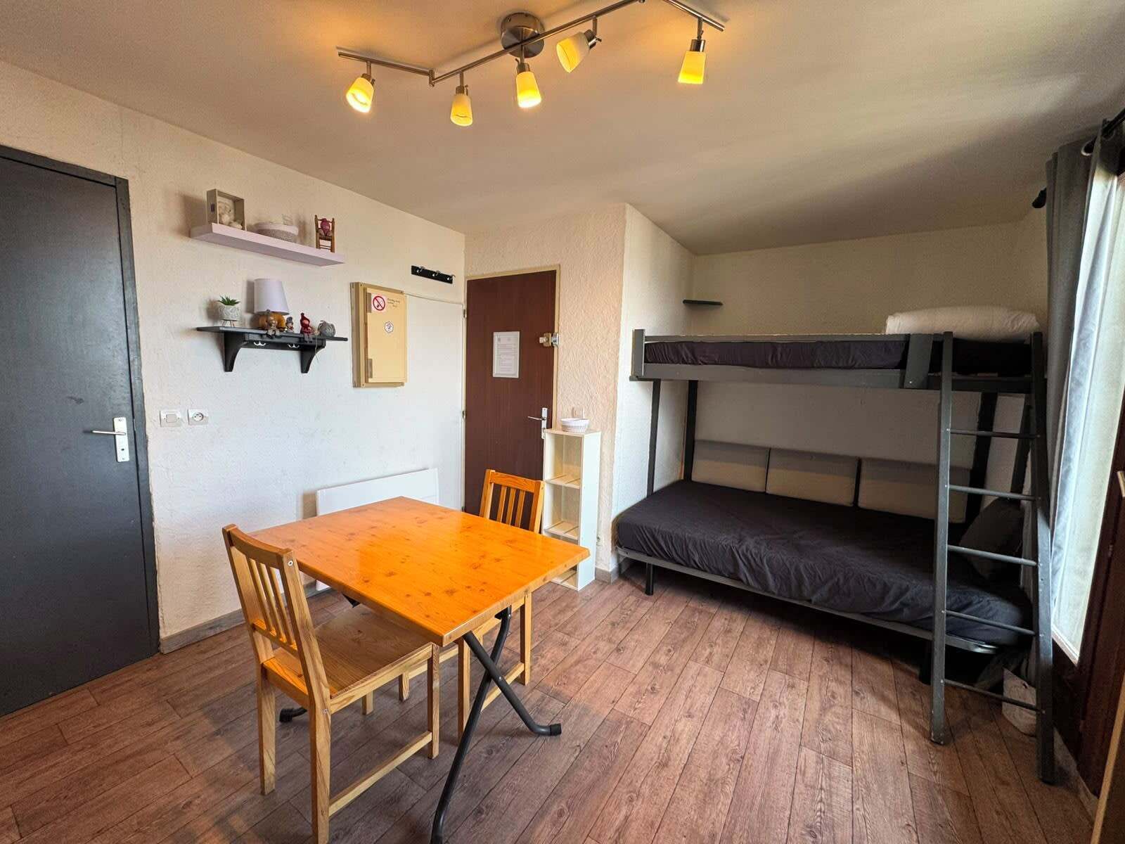 A compact room with a bunk bed, a wooden table with two chairs, a shelf with decorative items, and a door with a sign.