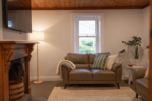 A living room with a brown leather sofa, a white throw blanket, a patterned pillow, a small side table with a plant, and a fireplace with a basket.
