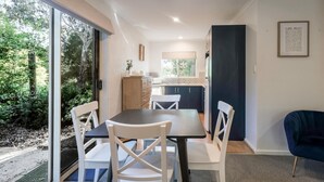 Un espace repas avec une table et des chaises, une cuisine en arrière-plan et une vue sur la verdure à l’extérieur.