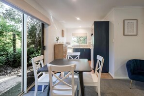 A dining area with a table and chairs, a kitchen in the background, and a view of greenery outside.