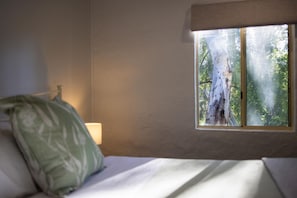 A room with a bed, a pillow, a nightstand with a lamp, and a window with a view of a tree and foliage.
