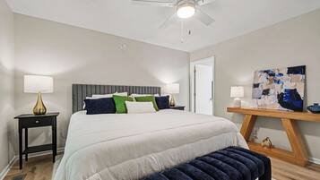 A bedroom with a bed, bedside tables, a bench, a ceiling fan, a painting, and a wooden floor.