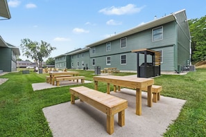 Un área de picnic con mesas y bancos de madera, un espacio de estar cubierto y un edificio verde al fondo.