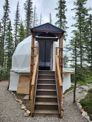 A wooden staircase leading to a covered entrance of a cabin-like structure surrounded by trees. - Riverside Buffalo Spirit Dome (Calgary)