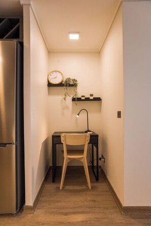 Eine kompakte Küche mit Kühlschrank, einem Holzbstuhl, einem Schreibtisch mit Lampe und einer Wanduhr.