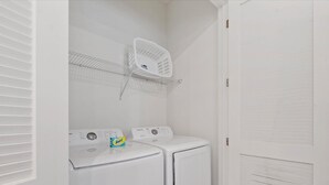 A laundry room with two white front-loading washing machines, a white shelf with a basket, and a white door with a vent. - Cozy 2-bedroom house 10 minutes to Disney! (Kissimmee)