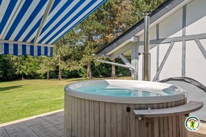 Ein Whirlpool unter einem gestreiften Sonnensegel, umgeben von einer Holzterrasse und einem Gebäude mit sichtbaren Holzbalken.