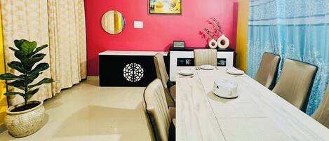 A dining area with a long table, chairs, a potted plant, a decorative wall piece, and a ceiling fan.