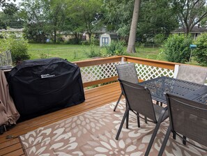 A wooden deck with a table and chairs, a barbecue grill, and a view of a backyard with a house and trees.