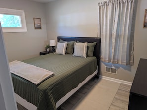 Una cama bien tendida con una colcha verde y almohadas, una mesita de noche con una lámpara y una planta pequeña, una ventana con vistas a árboles, y una cortina en la ventana.