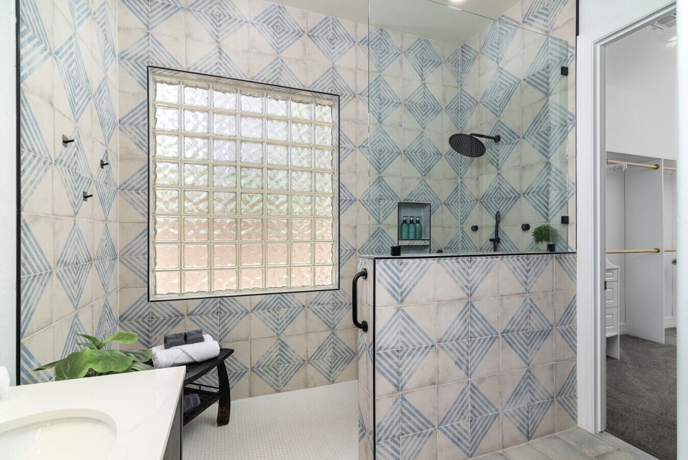 Une salle de bain avec un carrelage aux motifs géométriques, une fenêtre, une douche équipée d’un pommeau de douche à main et une petite plante posée sur une étagère.