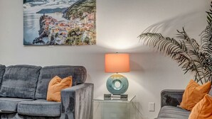 Ett vardagsrum med en grå soffa, ett soffbord i glas, en lampa med blå fot och en stor krukväxt.