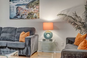 Ein Wohnzimmer mit grauem Sofa, einem Glastisch, einer Lampe mit blauer Basis und einer großen Topfpflanze.