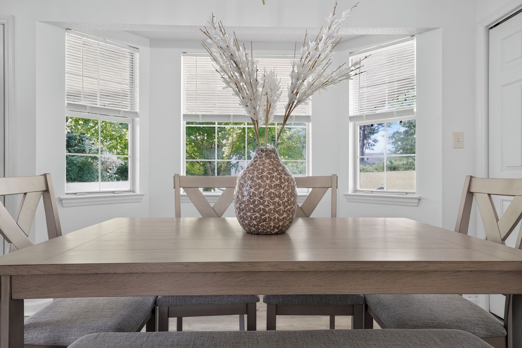 Una mesa de comedor con un jarrón decorativo, rodeada de sillas, en una habitación con ventanas grandes y paredes blancas.