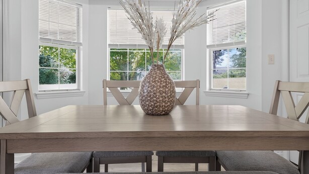 A dining table with a decorative vase, surrounded by chairs, in a room with large windows and white walls.