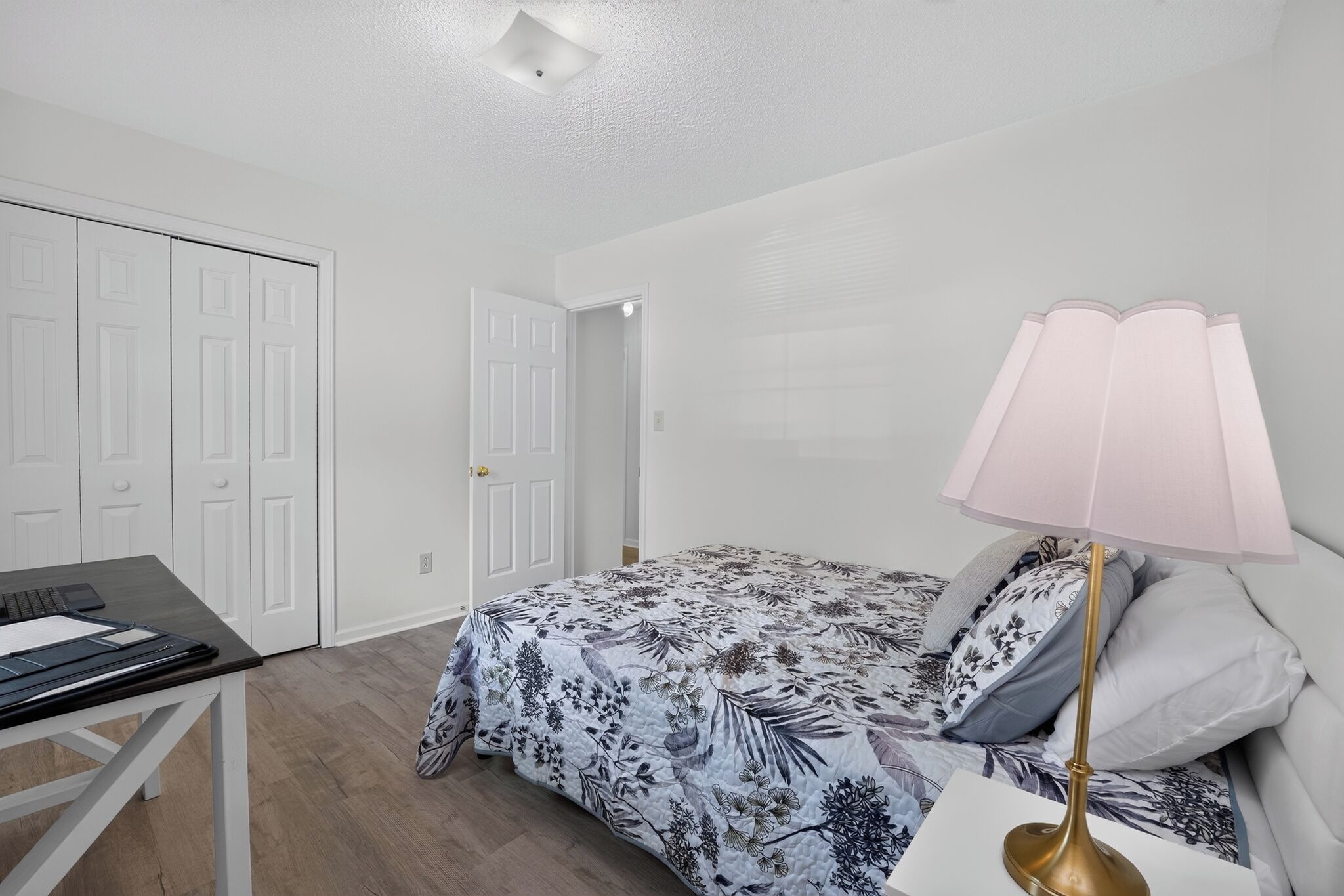 Un dormitorio con cama, escritorio, laptop, lámpara y puerta.