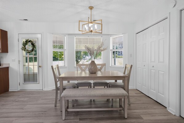 A dining room with a wooden table and benches, a chandelier, and a view through the windows.