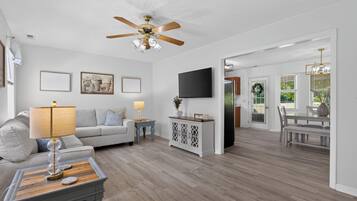 A modern living room with a ceiling fan, a flat-screen TV, and a dining area with a chandelier.
