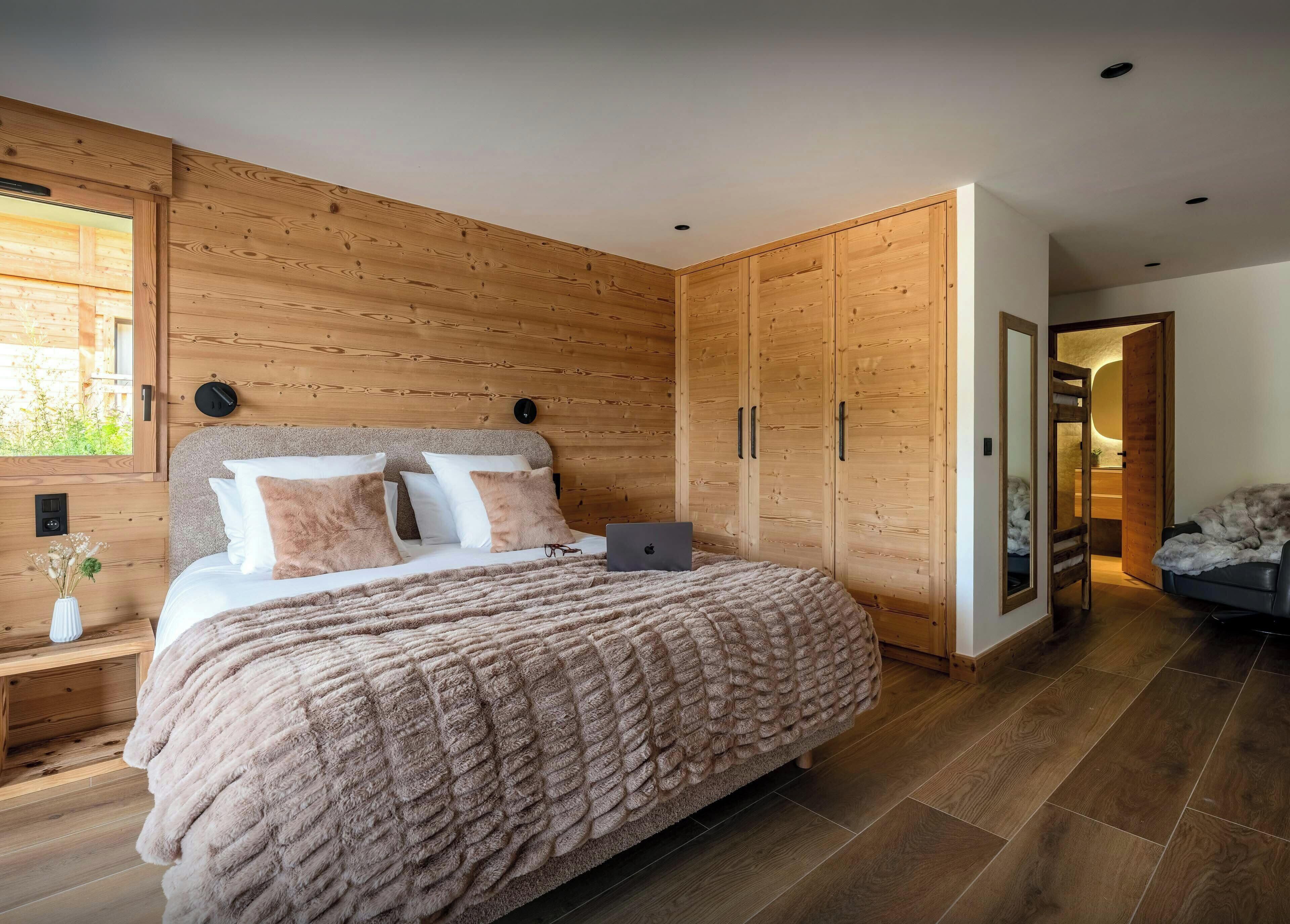 Un dormitorio con cabecera de madera, cama con manta suave, mesita de noche con un florero y armario con espejo.