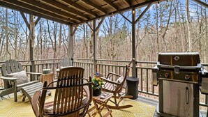 A covered outdoor deck with wooden furniture, a grill, and a view of a forest. - Long & Short of It - Pet Friendly, EV Charger, Firepit + Big Canoe Access (Jasper)