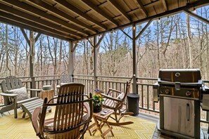 Eine überdachte Terrasse im Freien mit Holzmöbeln, einem Grill und Blick auf einen Wald.