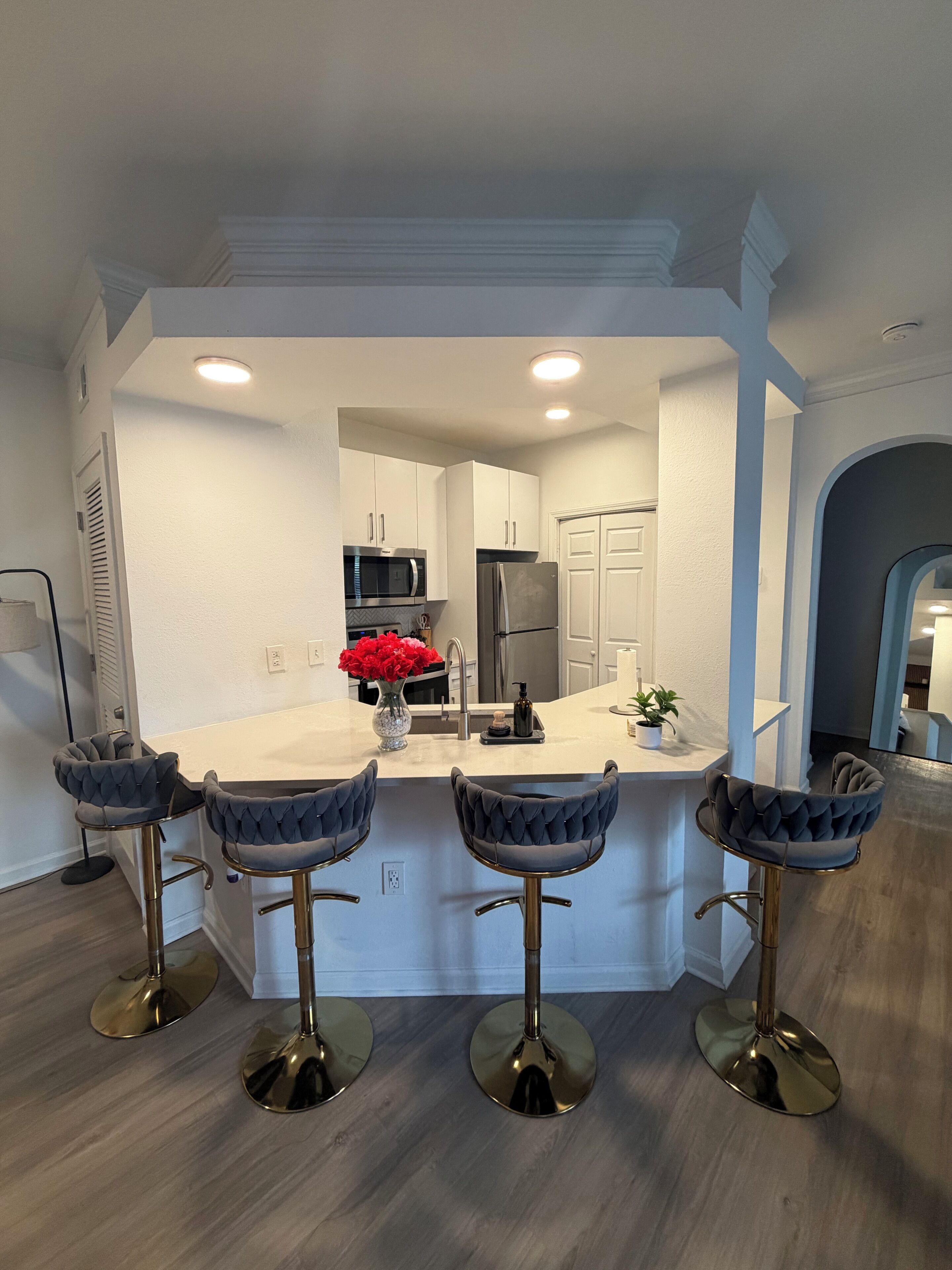 A modern kitchen with white cabinets, a stainless steel refrigerator, a microwave, and a sink. There are four bar stools with cushioned seats and gold bases. A vase with red flowers is on the counter.
