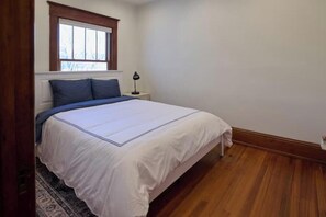 A bedroom with a bed, a nightstand, a lamp, a window, and a wooden floor.
