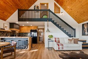 A spacious loft with a high ceiling, a central staircase, a kitchen with marble countertops, and a cozy seating area with a white sofa and a coffee table. - Painted Sky Cabin - Long Distance Mountain Views! | Firepit | Pet Friendly (Morganton)
