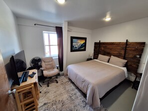 A bedroom with a bed, a desk with a computer, a chair, a window with curtains, and a wall-mounted picture.