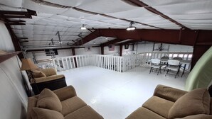 A spacious indoor area with a high ceiling, white walls, and a white floor. There are several brown upholstered chairs arranged in the foreground. - Country Retreat Barndo, great for groups, events, hunting camp & recreation. (Holbrook)