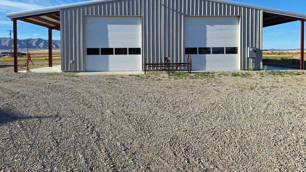 A single-story metal building with two large garage doors, a gravel driveway, and a bench. - Country Retreat Barndo, great for groups, events, hunting camp & recreation. (Holbrook)