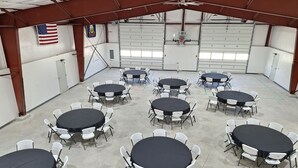 A large indoor space with multiple round tables and white chairs, a ceiling fan, and an American flag on the wall. - Country Retreat Barndo, great for groups, events, hunting camp & recreation. (Holbrook)