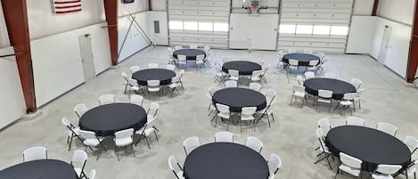 A large indoor space with multiple round tables and white chairs, a ceiling fan, and an American flag on the wall.