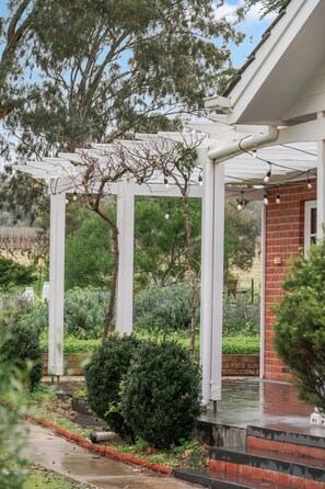 A covered outdoor area with a pergola, a brick building, and a garden with various plants.