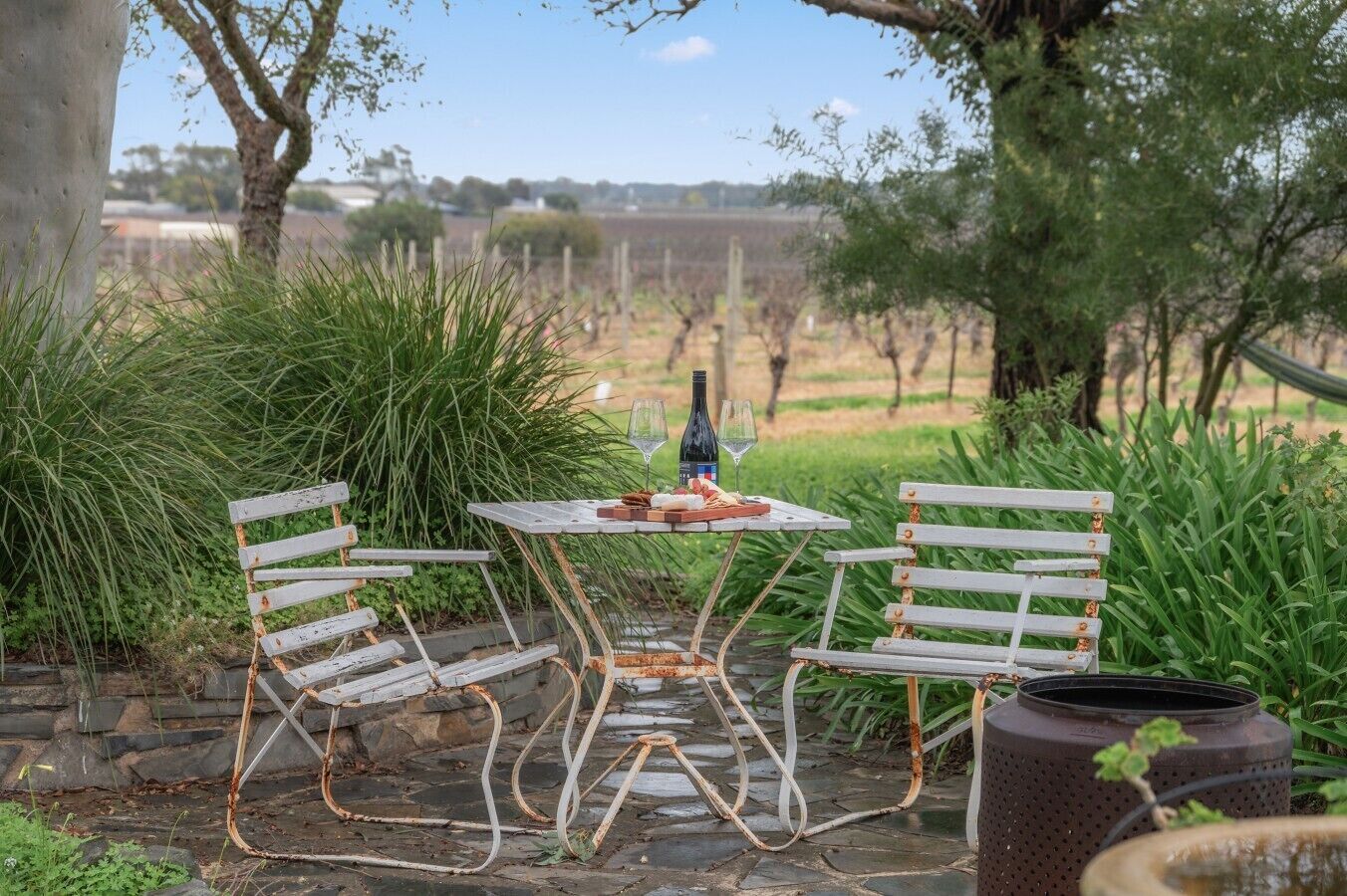 Un espace extérieur rustique aménagé avec une table et deux chaises, une bouteille et un verre, le tout entouré de verdure.