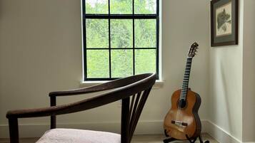 A room with a wooden chair, a guitar, a window with blinds, and framed pictures on the wall.