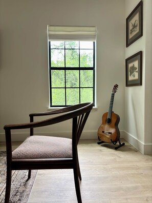 Una habitación con una silla de madera, una guitarra, una ventana con persianas e imágenes enmarcadas en la pared.