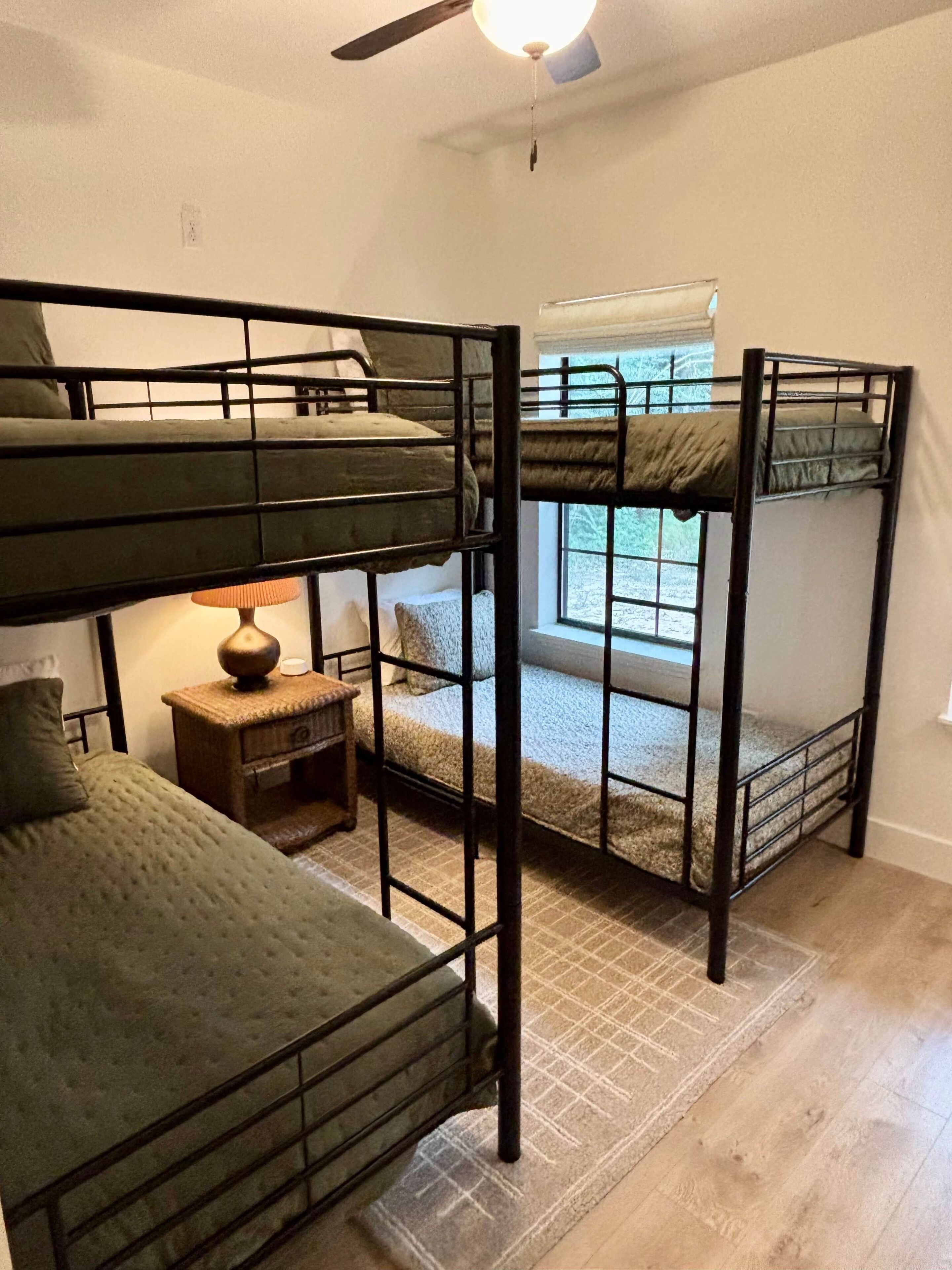 A bunk bed room with a ceiling fan, a small table with a lamp, and a window with a view.