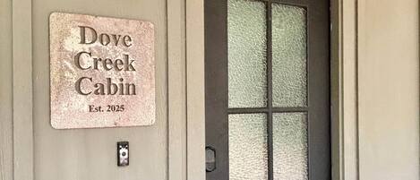 A door with a frosted glass panel, a metal doorbell, and a sign indicating 'Dove Creek Cabin Est. 2025' on the wall.
