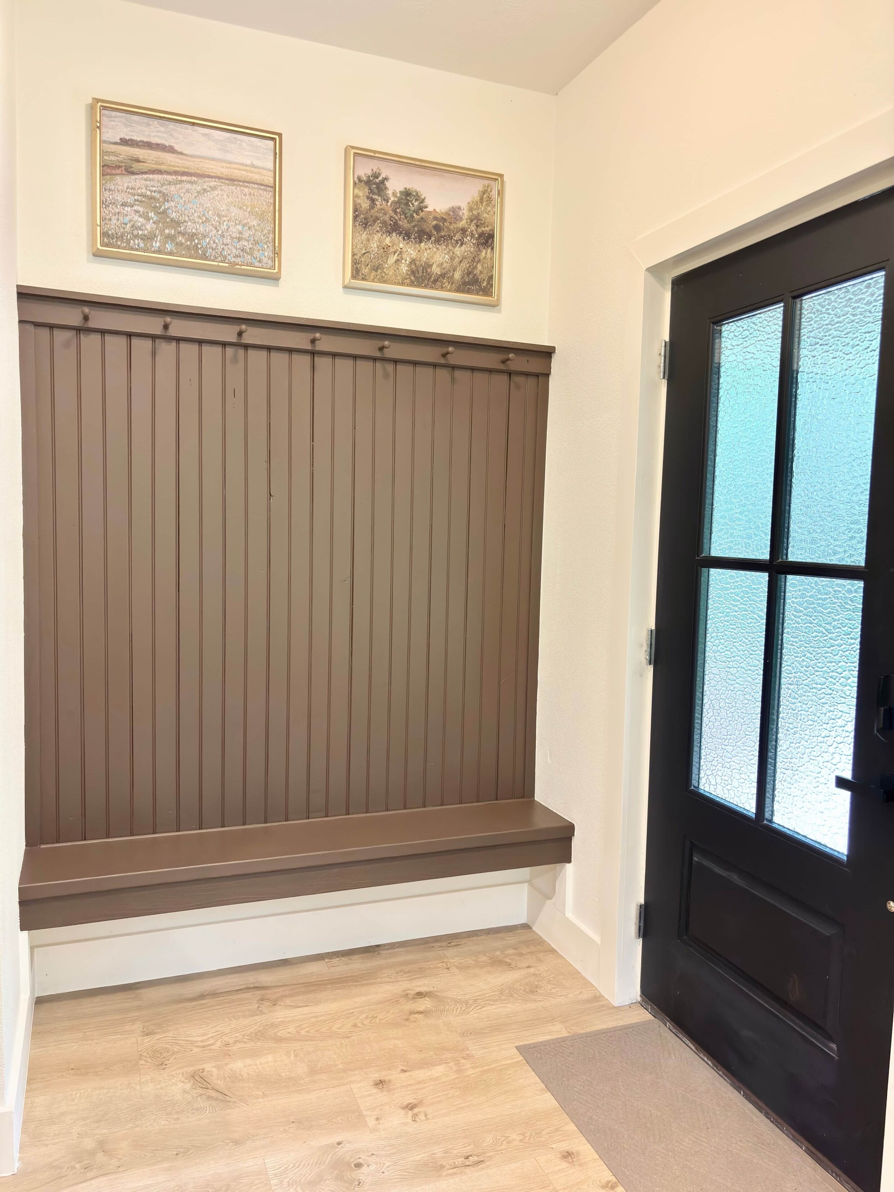Une pièce avec un banc en bois, deux tableaux encadrés au mur et une porte dont les panneaux sont en verre dépoli.