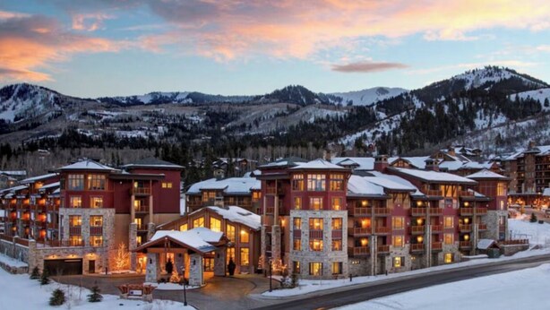 A multi-story lodge with a stone facade, large windows, and a snow-covered roof, surrounded by a mountainous landscape.