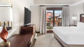 A hotel room with a bed, a television on a stand, a desk with a lamp, and a view of a building through a glass door.