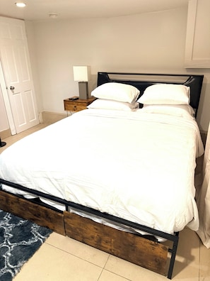 A neatly made bed with a wooden frame and white bedding, a bedside table with a lamp, and a door in the background. - StarBase Apartment (brooklyn)