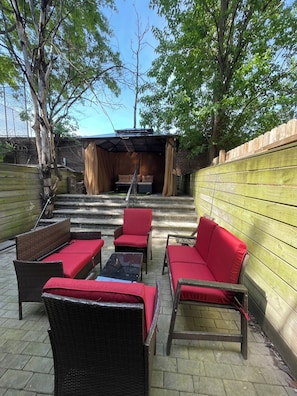 An outdoor patio area with wicker furniture and red cushions, a stone staircase leading to a covered seating area, and a wooden fence. - StarBase Apartment (brooklyn)
